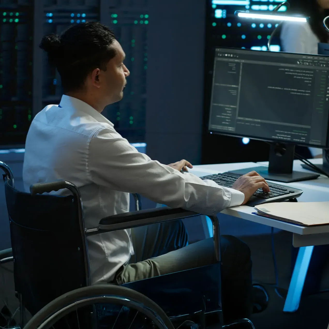 A focused cybersecurity professional working in a wheelchair at a desk in a high-tech server room, analyzing code on a large monitor, highlighting inclusivity and the practical skills emphasized in Herzing University’s graduate-level cybersecurity program.